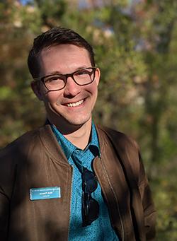 Nick smiles while standing in front of green foliage, in full sunlight